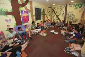 Students Sitting Along Walls of a Classroom with Tree Paintings and Signs Hanging Up